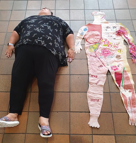 Life sized textile artwork of the body laying on the floor next to a woman laying down.