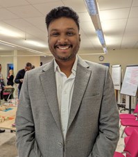 Brown skinned man wearing a grey suit jacket smiling at the camera