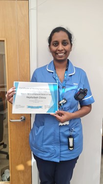 Woman wearing an NHS light blue top holding her certificate smiling at the camera.
