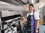 Lady wearing chefs clothes and an apron standing in a kitchen smiling at the camera.