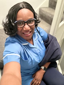 Black lady wearing glasses and an NHS light blue top, smiling at the camera