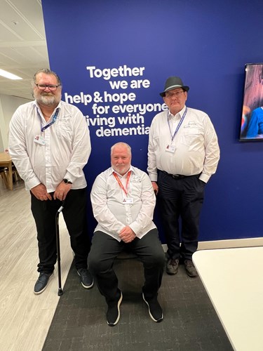 Three older men smiling at the camera. Two are standing and one is sitting.