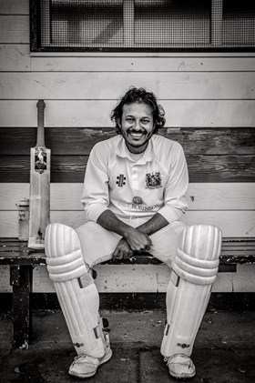 Black and white photo of a cricket player sitting down, smiling at the camera. 