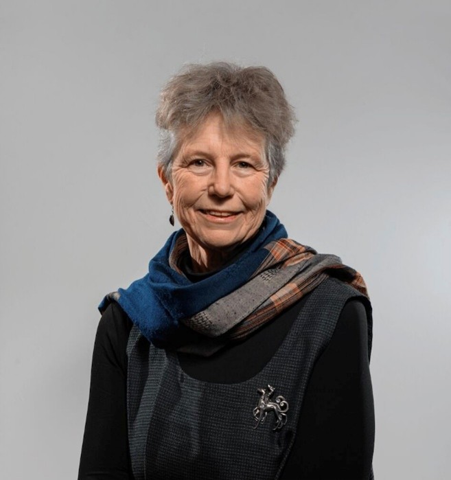 Elderly woman wearing a dark blue top, blue and patterned scarf around her neck and a brooch on her top.