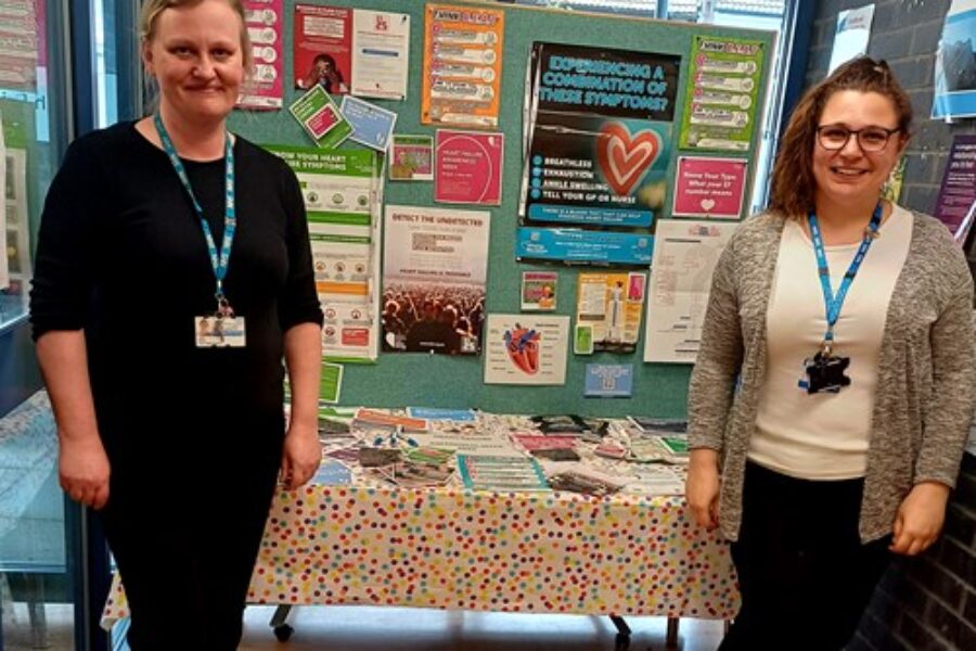 Two women smiling at the camera standing in front of the heart failure notice board.